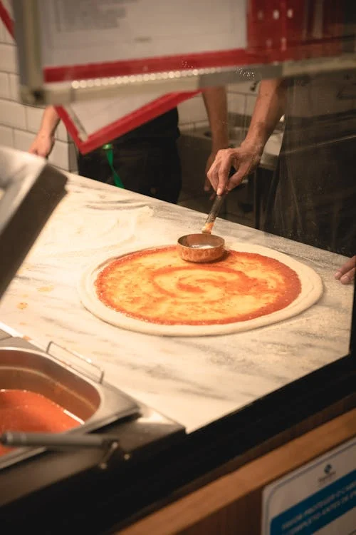 Pizza cruda e mano con un mestolo per spalmare la salsa di pomodoro sopra la pizza