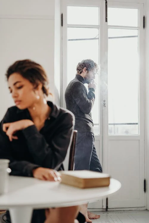 Foto di una ragazza seduta su una sedia e sullo sfondo un ragazzo che guarda fuori dalla finestra mentre fuma una sigaretta