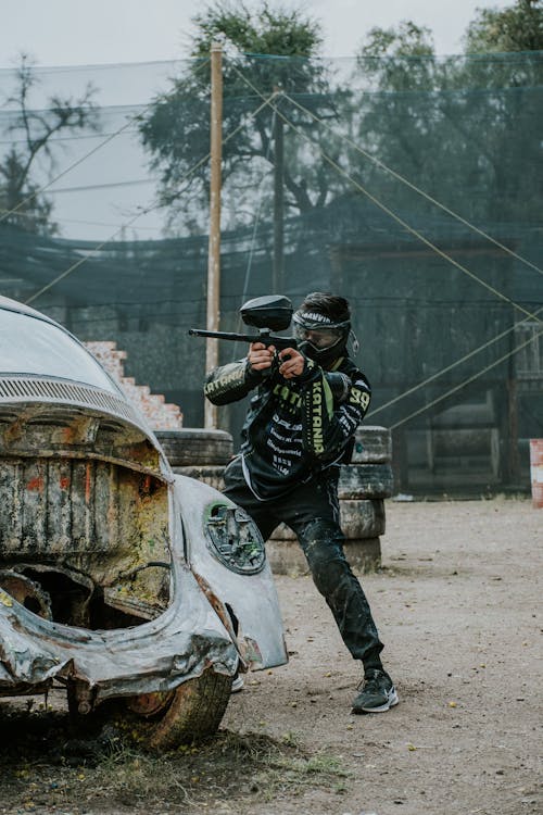 Foto di un ragazzo che sta giocando a paintball con un marcatore puntato che si nasconde dietro una vecchia macchina