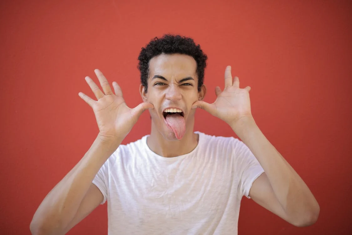 Foto di un ragazzo che fa delle smorfie con la lingua fuori e i pollici della mani sulle guance 