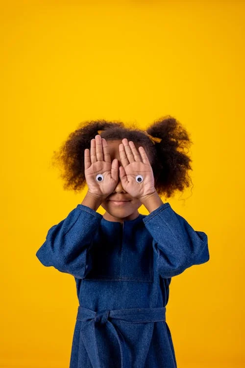 Foto di una bambina con le mani sugli occhi se degli occhi disegnati sulle mani, su sfondo giallo