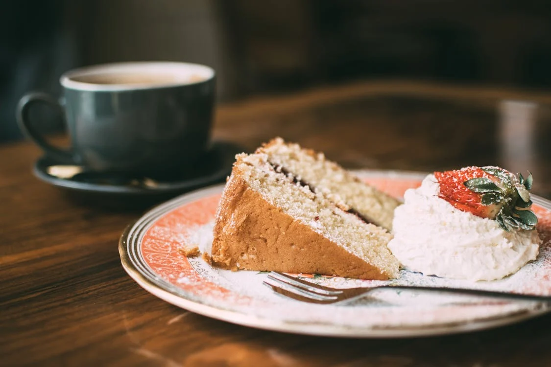 Foto di un piatto con dentro una torta e accanto della panna con sopra una fragola. Sullo sfondo una tazza