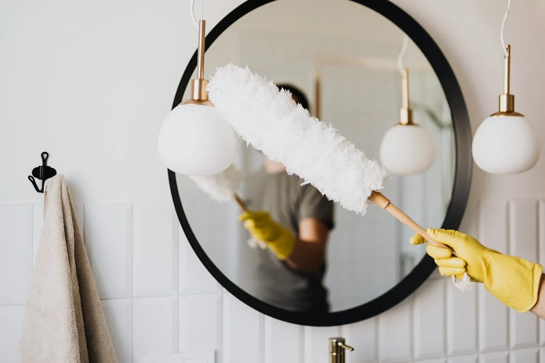 Foto di uno specchio e uno spolverino per pulire retto da una mano con un guanto giallo