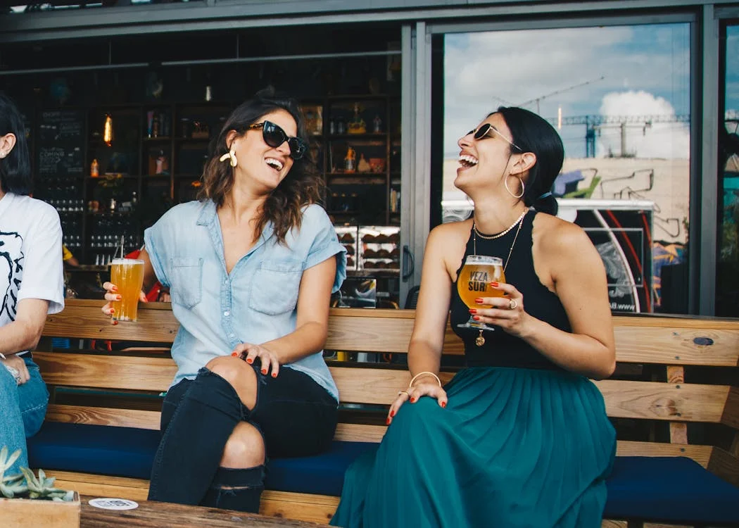Foto di due ragazze sedute su una panchina con della birra in mano mentre ridono