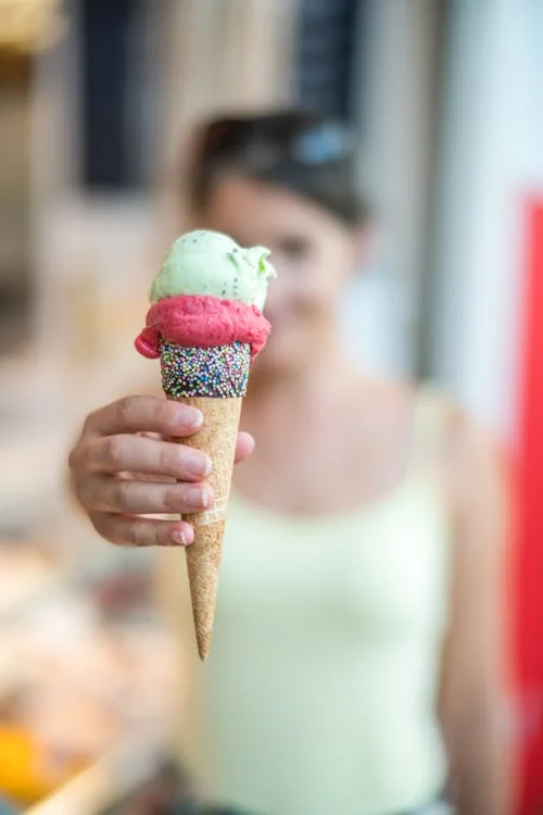 Foto di un cono di gelato in primo piano retto da una mano di una ragazza che si vede sfocata sullo sfondo