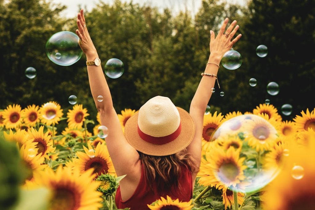 Foto di una ragazza di spalle con un cappello di paglia e le mani volte verso l'alto, in un campo di girasoli e delle bolle di sapone