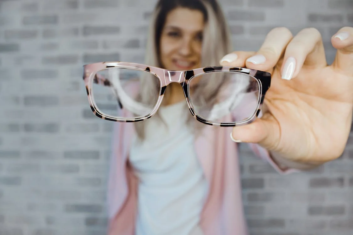 Foto di un paio di occhiali da vista in primo piano tenuti dalla mano di una ragazza che si vede sullo sfondo